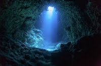 Distant underwater landscape with crystal clear water washing over dark cave walls. Light from outside shines through a hole in the ceiling, creating a pattern of turquoise rays that dance across the floor.