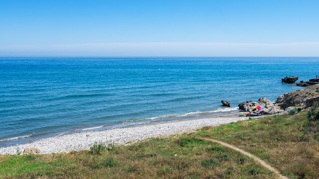 Pequeña cala con un camino que llega hasta ella entre la vegetación y el mar azul.