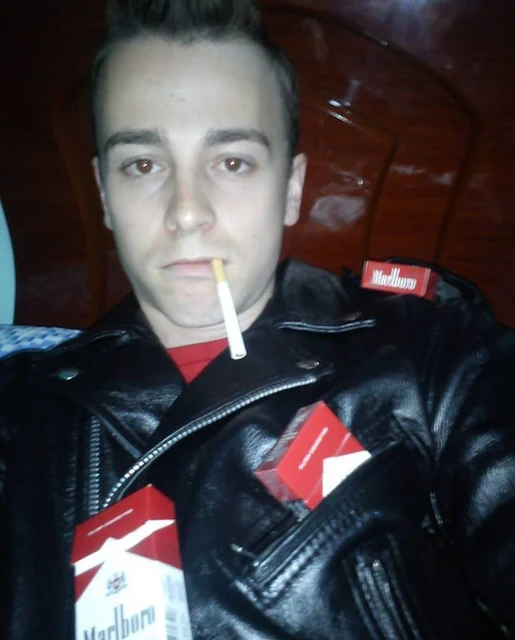 Shoulders up guy smoking a cigarette wearing a black leather biker jacket with a red stripe