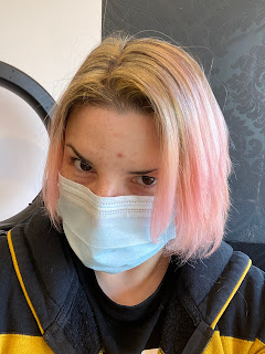 Lara, a pale skinned person staring into the camera. She is wearing a disposable face mask. Her hair is chin length with blonde streaks at the roots and washed out pink at the ends.