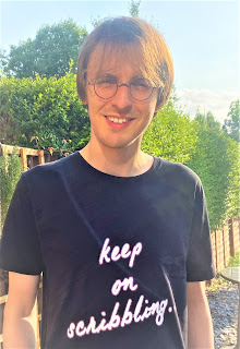 Photo of Richie Billing standing in a garden, facing the camera. Richie is wearing a black t-shirt with white text, outlined in pink, which reads "keep on scribbling." This is usually how he signs off his podcast, The Fantasy Writers' Toolshed.