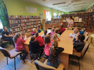 Bibliotekarka stoi na tle regałów z książkami i lady bibliotecznej.  Po lewej stronie  bibliotekarki znajduje się tablica z ilustracjami, obrazującymi zawód bibliotekarza o którym ona opowiada przed nią siedzącej na krzesełkach przy stolikach grupie słuchających ją dzieci z  Zespołu Przedszkolno-Szkolnego nr 3 w Szczakowej.