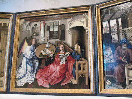 Medieval painting of the Annunciation. The Angel Gabriel in white on the left, the Virgin in red, reading a book, in the center. An ordinary room with a table and a bench in front of a fireplace, a window looking out on a landscape.