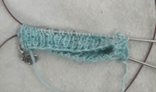 a small ribbed cuff being worked in fingering-weight yarn on circular needles using  magic loop. There is a small silver rose stitch marker clipped into the knitting.