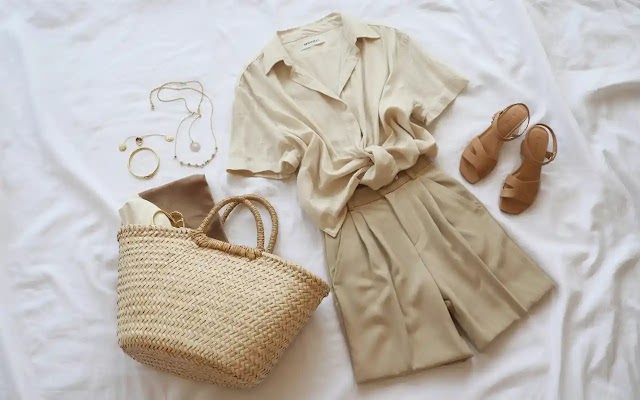 Flat lay of minimalist summer capsule wardrobe essentials for women including neutral linen blouse, beige trousers, straw tote bag, tan sandals, and gold jewelry on white linen background.