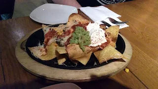 Oval wooden plate with cast iron plate on top of it. On the plate are a pile of triangular corn chips covered in shredded white cheese, yellow corn kernals, red tomato salsa, green guacamole, and white sour cream with red paprika sprinkled on top