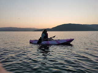 sunset on a loch. sky in shades of orange and yellow. woman in pink sea-kayak in middle ground on calm water.