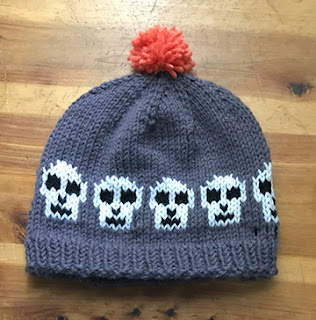 this is a picture of a grey hat with an orange pompom.  The hat has a row of white skulls with black features moving across hat just above the ribbing.