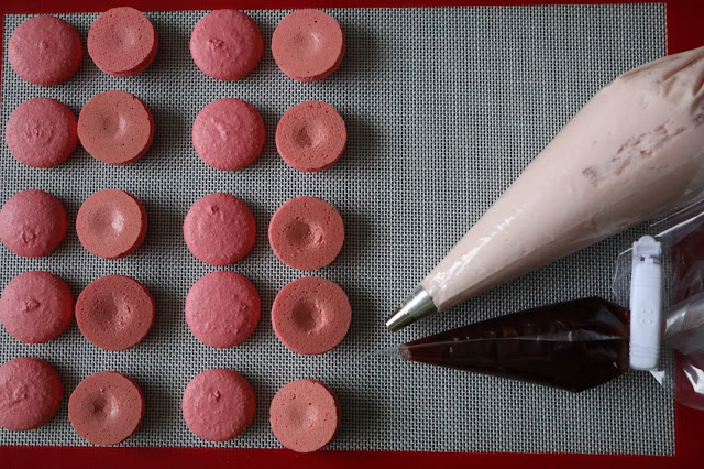 Pink Macaron Shells on a silicon mat with a piping bag filled with buttercream and another piping bag full of jelly.