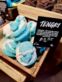 A group of blue spherical bath bombs with a geometrical design all over them in a large light brown wooden box with a black label with tenor in white font on a bright background 