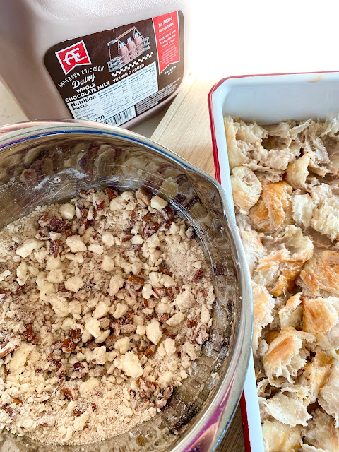 A white baking pan filled with croissant pieces, a mixing bowl full of streusel & AE Dairy chocolate milk in the background.