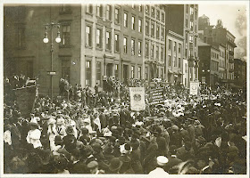 A black and white photograph of a parade moving through a crowded street.
