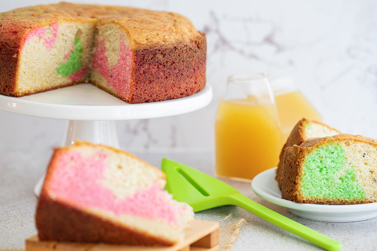 A plain sponge cake changed into a marble cake on a cake stand with a green spatula.