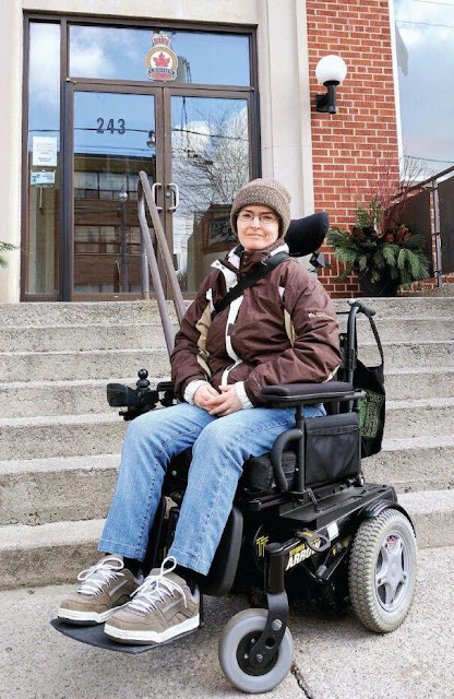 Image: a white woman in a winter coat and hat sits in a power wheelchair in front of an inaccessible set of stone steps