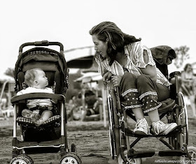 Baby carriage next to a wheelchair, both viewed from the front, baby in carriage, young adult woman in wheelchair, turned to her left to talk to the baby, with the baby looking at her