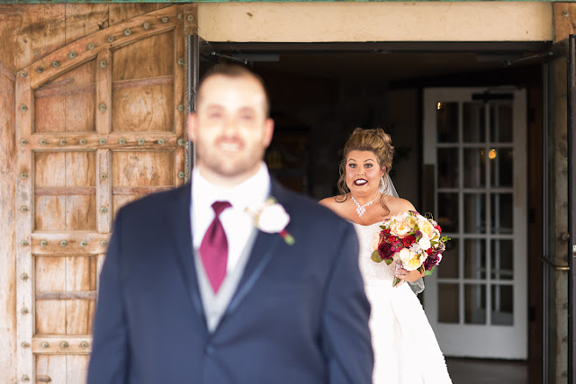  Cassie and Wyatt tied the knot at The Palace Event Center, in Yukon, OK, on July 15, 2017.   The venue’s magnificent wood entry doorways, outside rock façade, and adjacent country landscape provided the perfect backdrop to photograph the first look and pictures of the jovial wedding party.  The venue was spacious, which allowed us to sneak in a few pre-dressed shots of the girls in their “Wedding Tribe” t-shirts.     Cassie’s elegant, form-fitting, strapless, satin wedding dress overlaid with lace and sequins looked like it was made especially for her.  Wyatt was quite the Knight in Shining Armor dressed in his navy suit, grey vest, and burgundy tie.  The four beautiful bridesmaids wore matching full-length, flowing, burgundy gowns, slit at the knee.  Cassie’s gorgeous bouquet was composed of baby’s breath, peonies and ranunculus in rich hues of vibrant burgundy, red, pink, burnt orange, and yellow gold.  To add to the excitement of the day, their son had a special assignment—carrying the sign down the aisle that stated, “Daddy, Here Comes Our Girl.”     The venue’s grand ballroom, with it’s beautiful chandeliers and romantic lighting, was the perfect backdrop for the couple’s reception.  Candlelight, flowers, and twinkle lights adorned the nuptial stage, and after the vow exchange, Cassie and Wyatt participated in their first dance.  Guests were invited to enjoy the buffet, the cutting of the cake, and the toasts by the best man and bridesmaid.  Chairs were quickly removed from the dance floor, and while almost everyone spent the rest of the afternoon relaxing, dancing and socializing, there was a particularly fun-loving group of guests who decorated the bride and groom’s Jeep with sayings, crepe paper, and a car filled-to-the-brim with balloons!        Cassie stated that the best part of the wedding was being able to see so many family and friends at the reception having fun and enjoying themselves.  She said, “If we had it to do over, we wouldn’t do anything differently…it was perfect for both of us!”     It was a pleasure to capture the special moments of this memorable wedding day for Cassie and Wyatt, and we wish them a lifetime of love and happiness. 