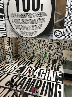 photo of the Barbara Kruger installation at the MOMA - text covers the floor and walls, towering over the people in the space