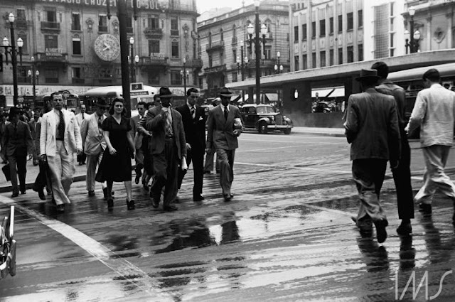 A fotografia negativa mostra pedestres atravessando uma rua. O chão está molhado. Os homens usam terno e gravata, e a única mulher visível está de vestido e sapato de salto. Os prédios não possuem mais do que cinco andares.