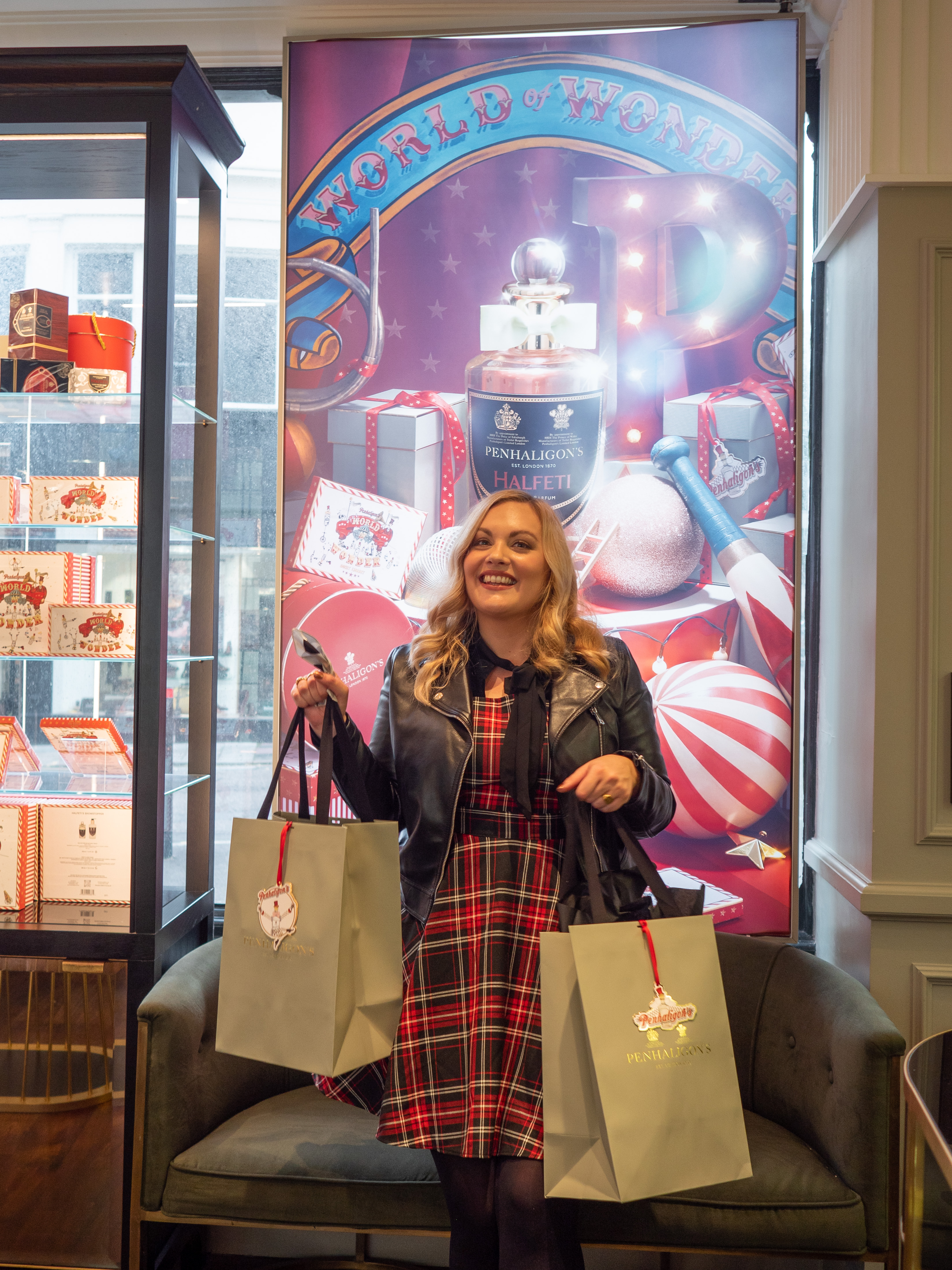 Penhaligon's Store Brighton: World of Wonder Christmas Collection 2022. Blogger Katie holds bags of Penhaligons goodies inside the store in front of a big Halfeti Christmas poster. She wears a red and black pinafore dress and black leather jacket.