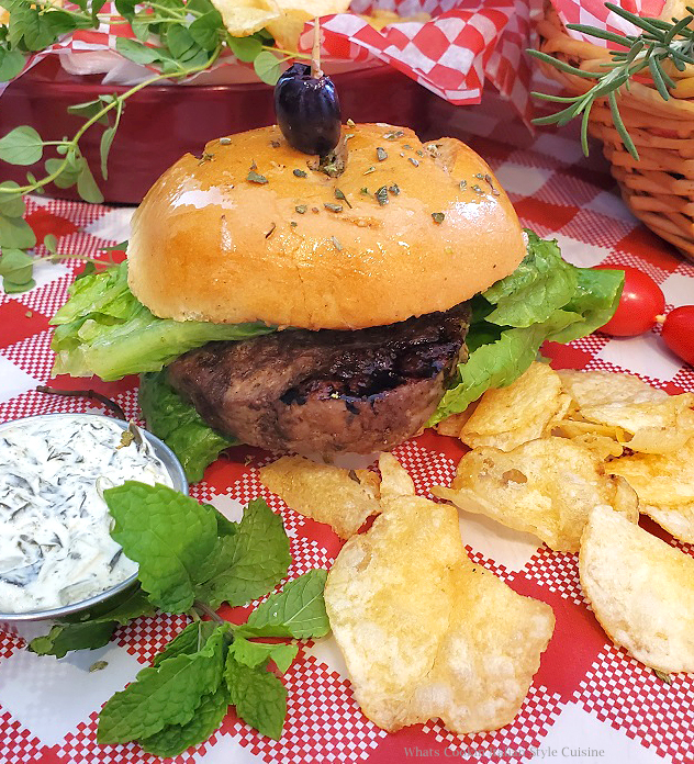 this is a lamb burger on a red checkerboard tablecloth along with potato chips and  with spinach tzatziki sauce