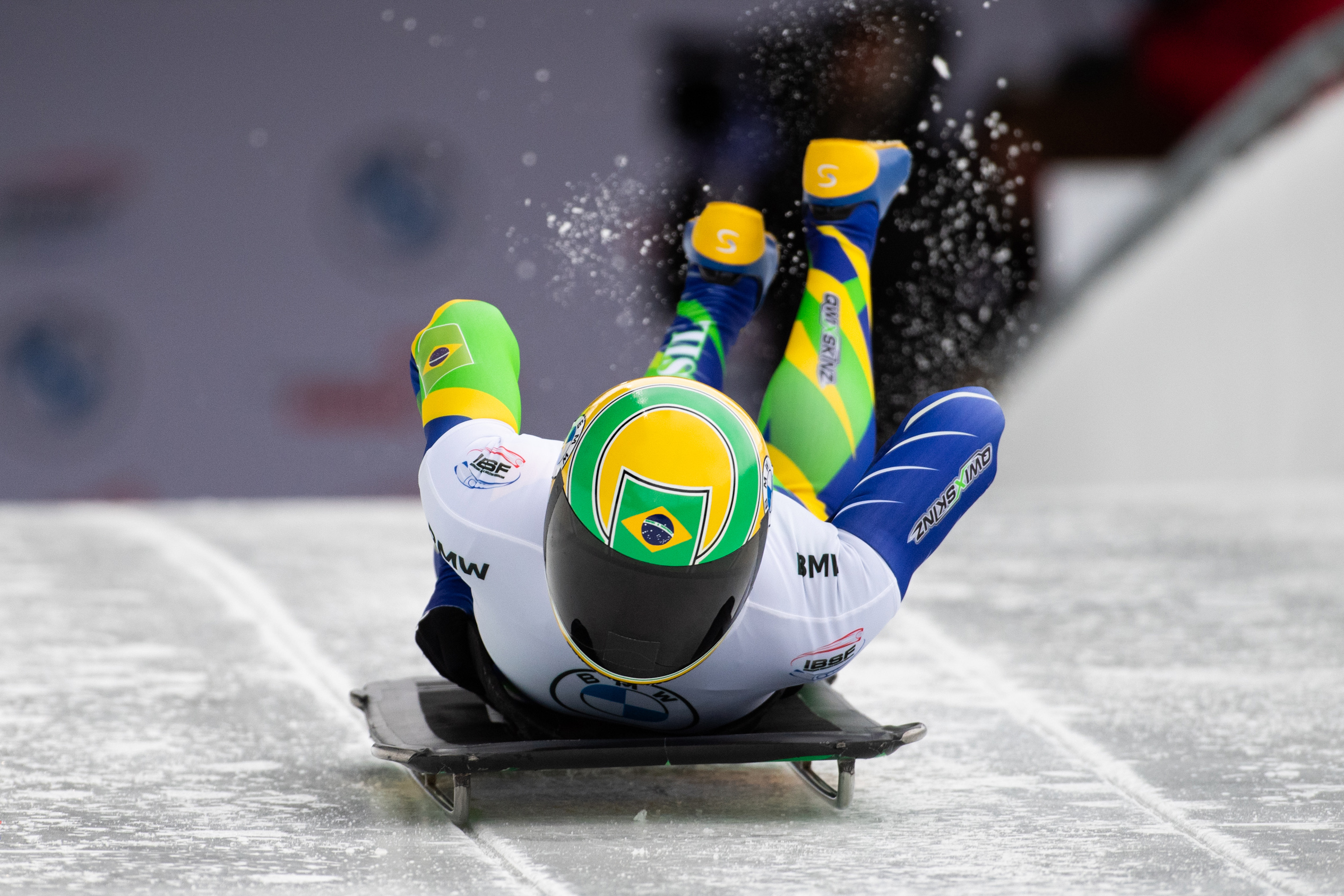 Nicole Silveiar, com colete branco em cima de uma roupa azul e com capacete amarelo com as cores do Brasil, desce uma pista de gelo de bruços em um trenó