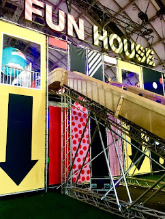 A stand containing a bright stone coloured slide with different coloured steps and yellow arrows on the side with FUN HOUSE in bold silver letters above on a bright background.