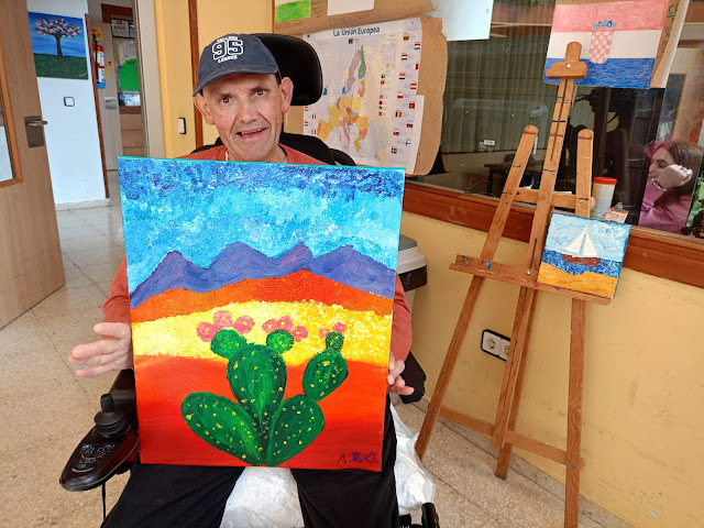 Un hombre sosteniendo un cuadro que ha terminado, en el que se ve un cactus con flores y al fondo unas montañas