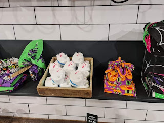 A landscape photo of a light brown square box filled with white ghost shaped bath bombs with red, yellow, blue and purple glitter next to a green plant next to a white and black bat print drawstring bag on a light brown rectangular shelf on a bright background