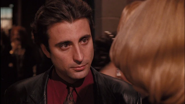 Shoulders upward Andy Garcia talks to blonde wearing black leather blazer with red shirt and gold tie underneath