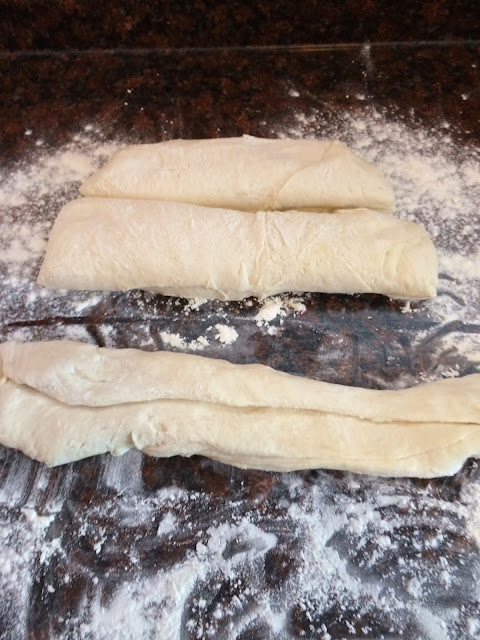 Flour on the counter with the three dough portions.  One is being flattened out and rolled into a snake.