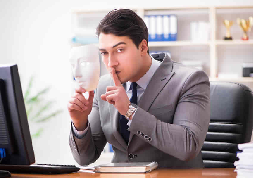 Imagem mostra homem de terno sentado de frente para uma mesa onde tem um computado e uma agenda com uma das mãos ele faz sinal de silencio com a outra segura uma mascara branca