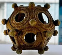 Bronze dodecahedron with 12 holes located symmetrically over the entire surface. The ball lies on a well-lit wooden table. Its surface is smooth, slightly patinated.