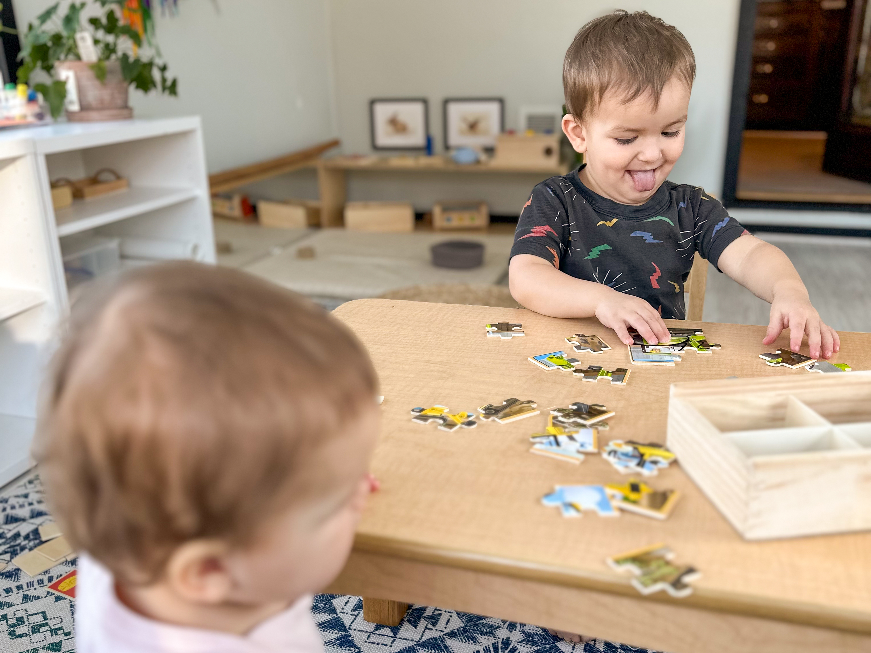 Toddler happily works on jigsaw puzzle at table in Montessori home while baby watches on.