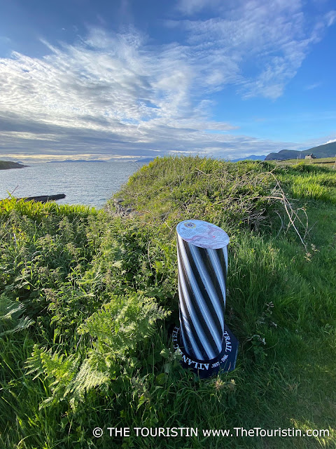 A black and white pillar/monument of a transatlantic cable in the midst of a green meadow by the ocean in soft evening light.