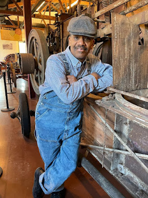 An African American man with a mustache and wearing denim overalls, a blue and white check long-sleeved shirt, and a gray tweed cap leans against a piece of antique farm machinery. He is portraying inventor William Chester Ruth, and the machinery is inside an exhibit gallery.