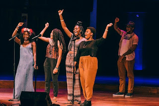 Photo of the 2016 DC Youth Slam Team on stage at the 2016 Brave New Voices International Youth Poetry Slam Festival in DC. The four young women are smiling with one hand raised in a fist as they share their team chant. The Youth Programs coordinator, Joseph Green, is in the background with his hand up too.