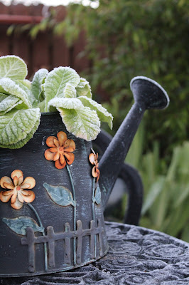 A dark green watering can shaped flowerpot, decorated with orange flowers. In it is a plant with no flowers, just light green leaves, covered in a thin layer of frost.