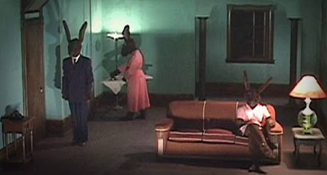 Three human sized rabbits in a living room setting. One is stood by the door, one is doing the ironing in the left corner and one is sat on the sofa.