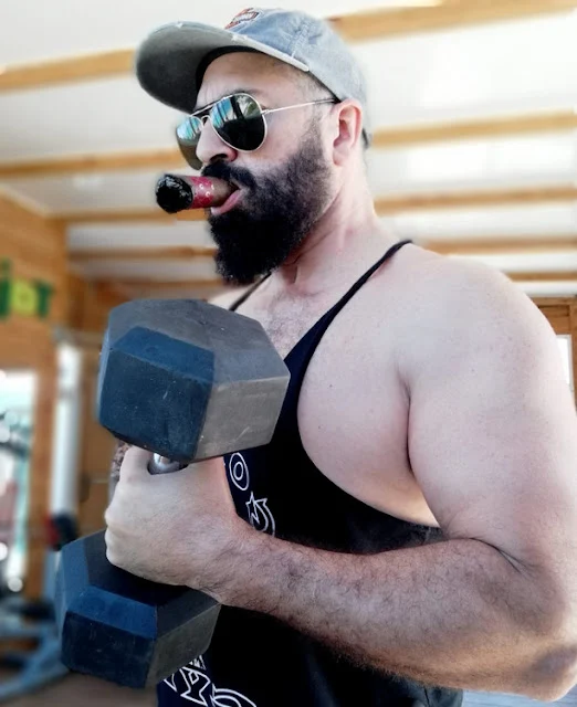 E 2/2 Muscular man with a large bushy dark beard wearing sunglasses and the muscle shirt lifting weights outside with a cigar in his mouth
