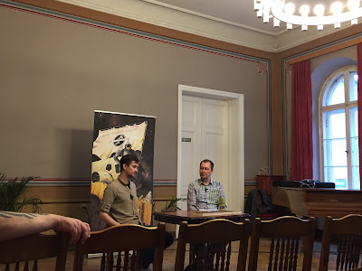 Hetk raamatuesitluselt Tartu kirjanduse maja saalis. Noor autor Joonas Kollo ja vana ulmik Veiko Belials vestlevad romaanist "Tilk tõrva tulevikku". Kaadrisse on jäänud ka pealtvaataja käsi.