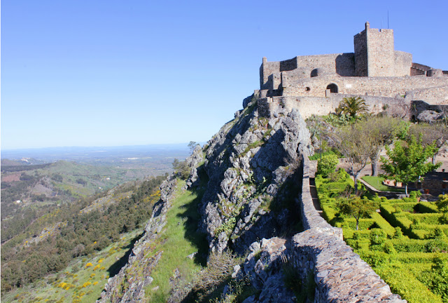 Średniowieczny zamek w Marvao. Na skraju skarpy ciągnie się mur miejski. Po jego stronie wewnętrznej znajduje się ogród o regularnie przystrzyżonym żywopłocie. Po stronie zewnętrznej muru jeżą się skały, za którymi otwiera się szeroki widok na równinę. Mur dobiega do budunku zamku - piętrzących się jeden wyżej drugiego prostych, szarych murów, ponad które wystaje kwadratowa wieża. 