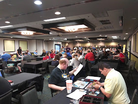 A view of the gaming hall. A long room full of tables, with lots of people at those tables playing a variety of games. At the far end, the games library can be seen.