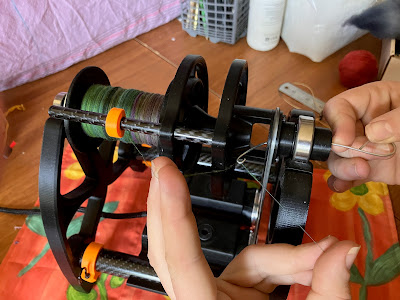 The Falcon e-spinner in the process of threading the orifice hook. One hand is holding the orifice hook, which is threaded through the orifice. The other hand has pinched a length of spun thread taut between index and middle fingers close to the bobbin, and thumb and ring fingers near the orifice. The orifice hook is wrapped around the taut thread, ready to draw it through the orifice.