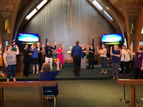 people in a chancel, in a line, signing together, with a leader in front and screens behind