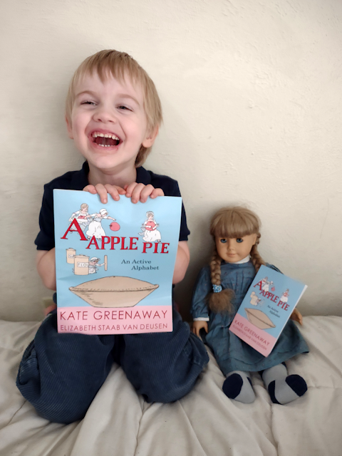 A laughing small boy kneels next to an 18" doll as a size comparison. The boy is holding a large 8.5x11 book and the doll is holding a small 4x6 book.