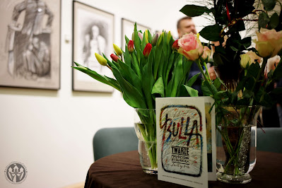 zdjęcie z kwiatami w wazonie i folderem z wystawy Ignacego Bulli. W tle obrazy artysty
