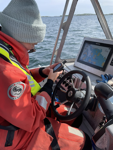 Henkilö katsoo käsi-GPS-laitetta ajaessaan veneellä, päällä pipo ja pelastautumispuku.