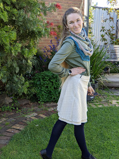 The author poses for the camera in the backyard: her right hand is on her hip and she is turned half away from the camera to show the side of the skirt. She wears a handknit shawl, a green shirt, the cream skirt that is the subject of this post, navy tights, and black Oxford shoes.
