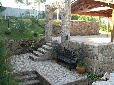 Construção de muro de pedra bruta com dois tipos de assentamento de pedra. O muro de pedra faceado e o muro de pedra solta sem cimento com nichos para colocação de plantas com as escadas de pedra bruta, o piso de pedra bruta, o pórtico de pedra e a execução do paisagismo com os vasos de plantas e o banco de madeira.