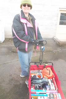 Cynthia M. Parkhill stands holding handle of a wagon, shown from rear, top-down-view. The wagon is near-full of books, an orange drawstring bag, bike lock and pair of gloves